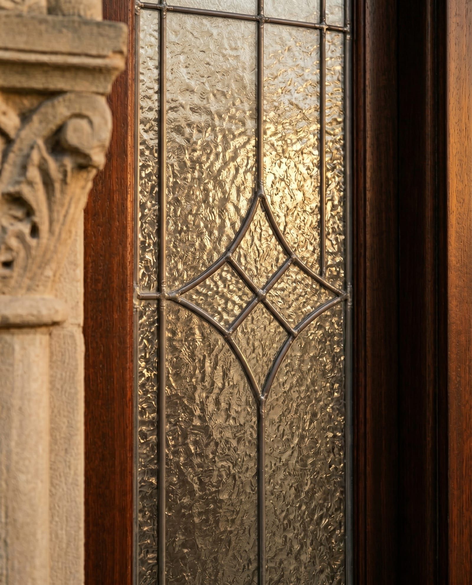 Close-up of a leaded-glass sidelite with a clear beveled diamond medallion centered on a textured cathedral glass field, in a dark mahogany casing next to a carved limestone column
