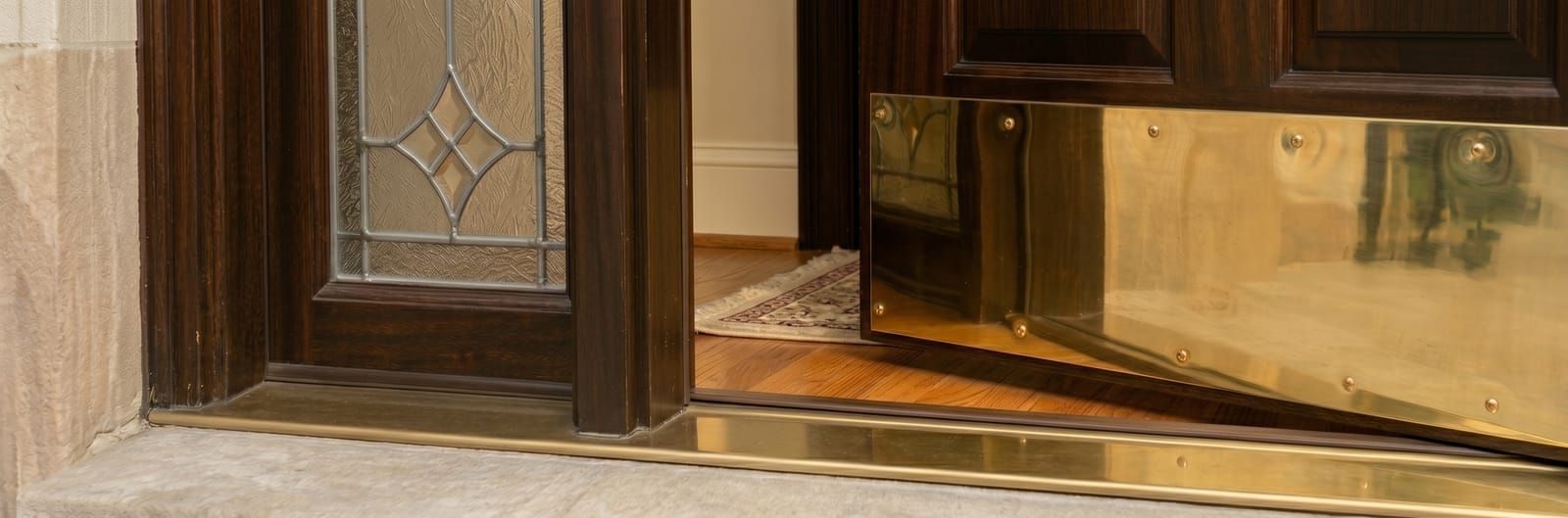 Close-up of the open mahogany door's threshold — polished brass kickplate on the slab, brass sill saddle on a limestone step, leaded-glass sidelite to the left, oak hardwood floor visible inside