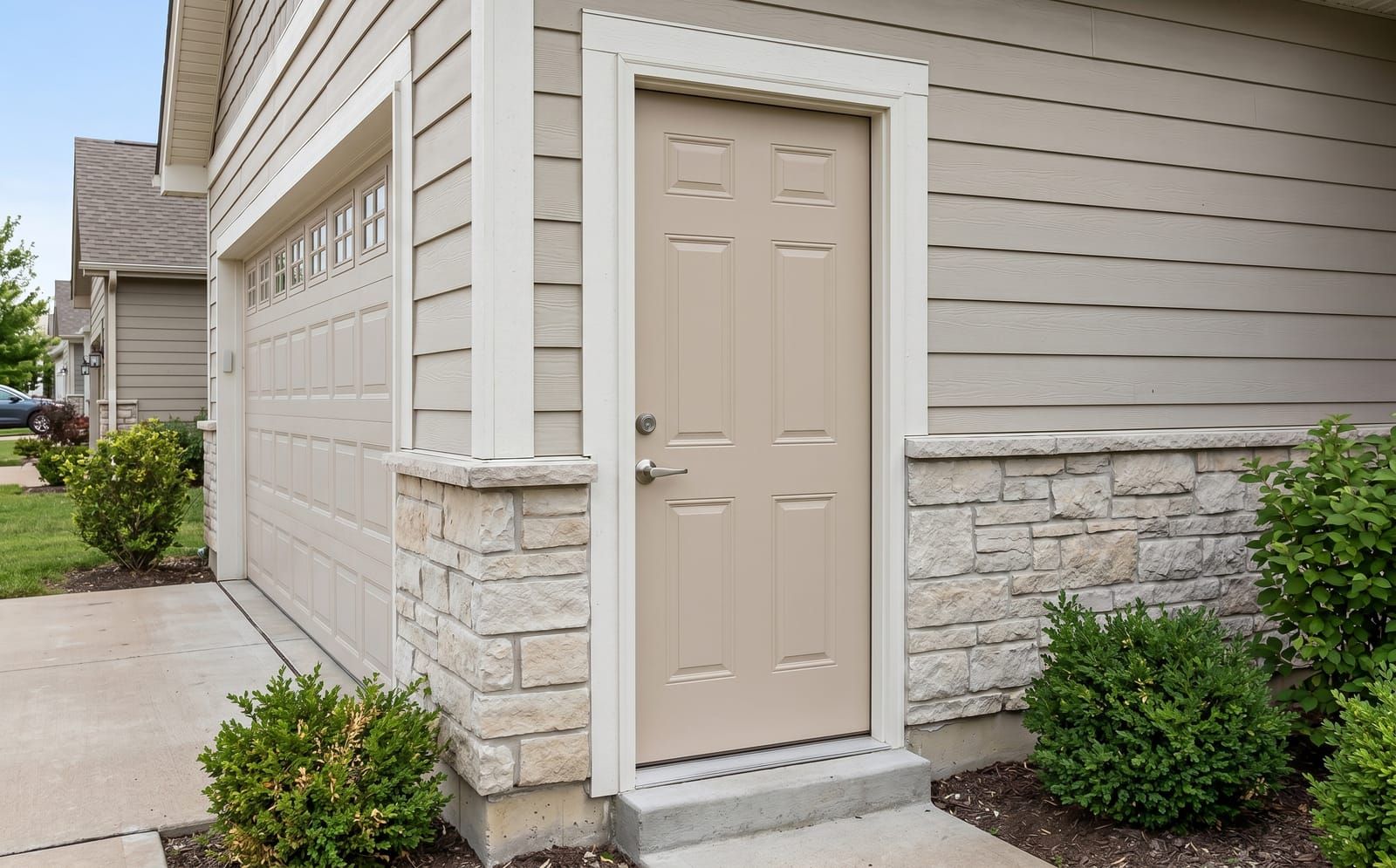 A beige 6-panel residential steel exterior door installed as the side pedestrian door of an attached garage with stone wainscot and lap siding