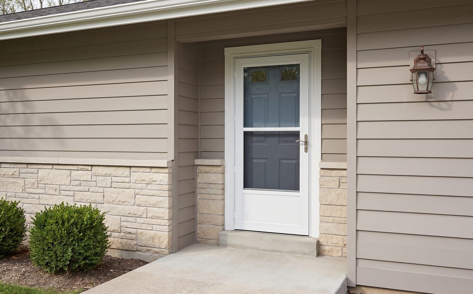 White-frame mid-view storm door with kick-panel and self-storing glass installed over a slate-blue main entry door on a Hardie-board ranch home with stone wainscot