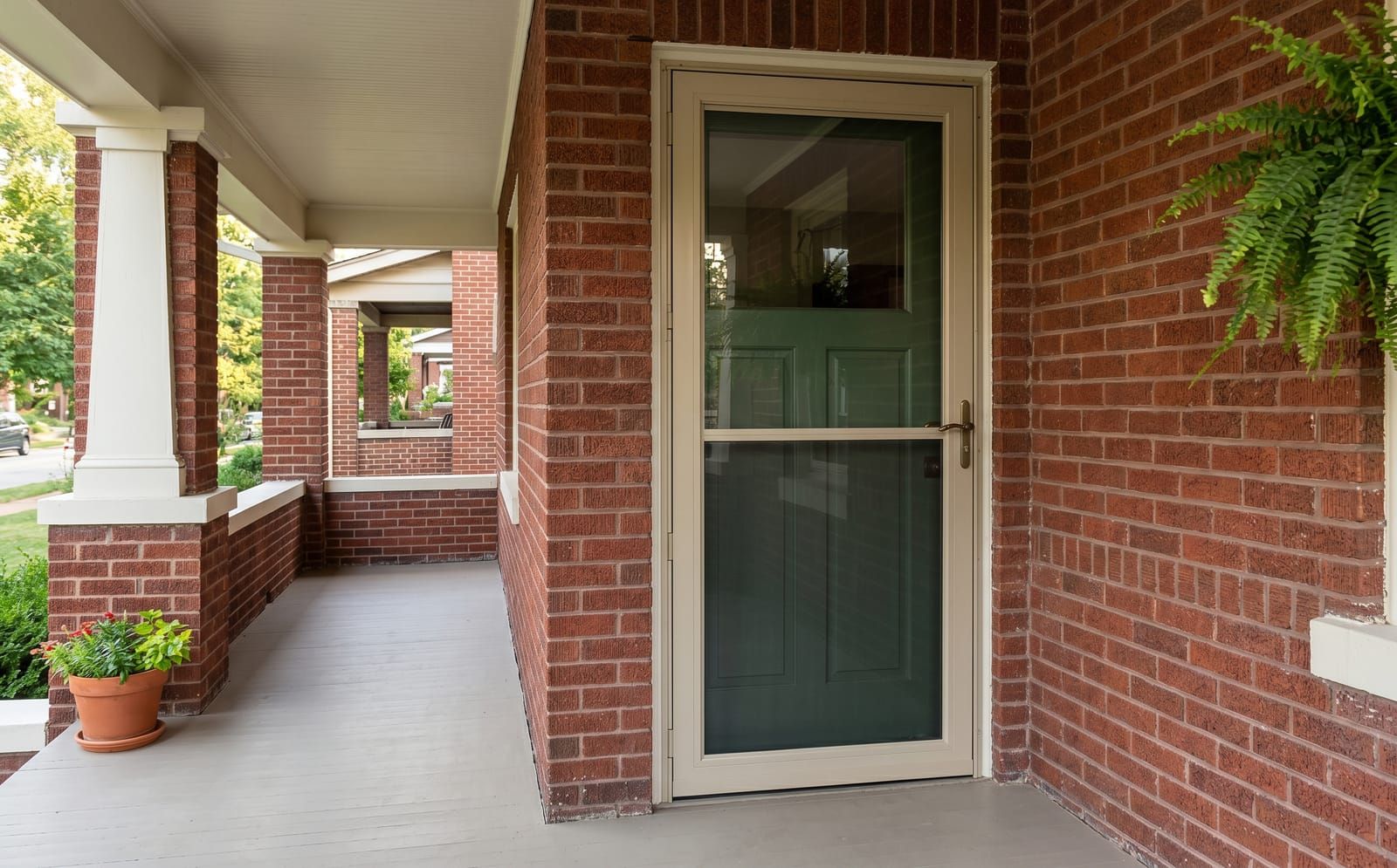 Sandstone aluminum-frame storm door with hidden retractable screen, installed over a forest-green main door on a brick craftsman porch